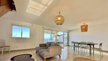 A spacious living area with a sofa, dining table, and chairs, large windows, and a view of the sea.