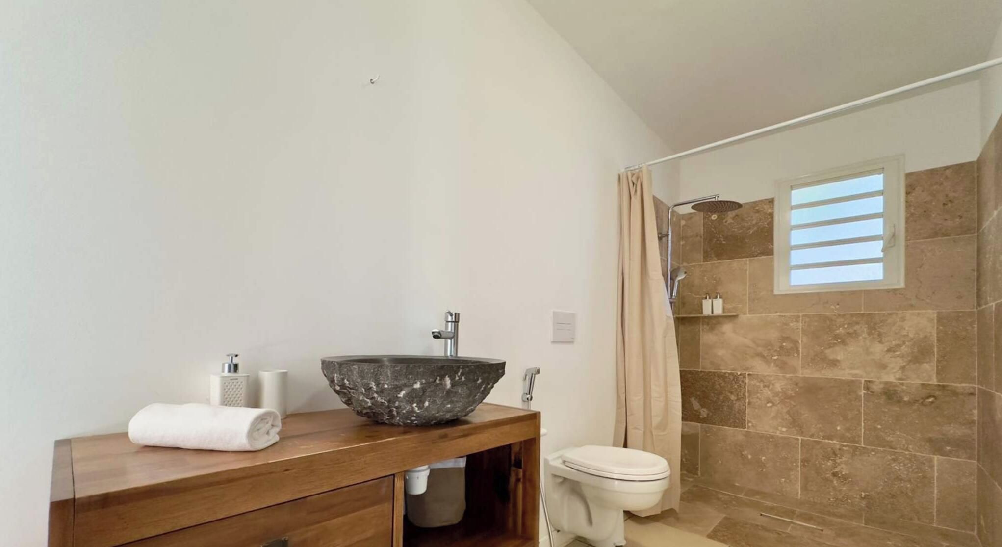 Une salle de bain avec un lavabo en pierre, un meuble-lavabo en bois, des toilettes et un espace douche avec une fenêtre.