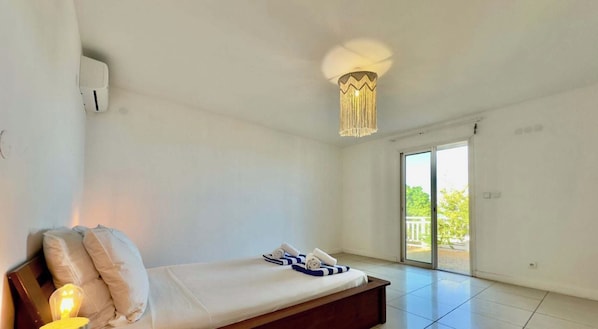A room with a bed, a bedside lamp, a ceiling light fixture, and a sliding glass door leading to an outdoor area. - Whale Beach Villa by HILO Collection (Les Trois-Bassins)