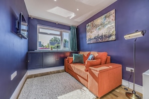 A room with a dark blue wall, a window with curtains, a sofa with cushions, a painting, and a floor lamp. - Charming House with Pretty Garden & Parking - Pass the Keys (Chichester)