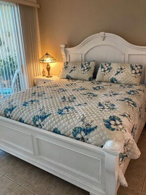 Una cama con colcha floral, una mesita de noche con lámpara y vistas a un balcón.