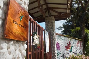 A wooden sign with 'Villa Cristina' and 'Casa Boutique' text, a stone wall with a floral mural, and a metal gate. - Villa Cristina - Casa Boutique (PEREIRA)