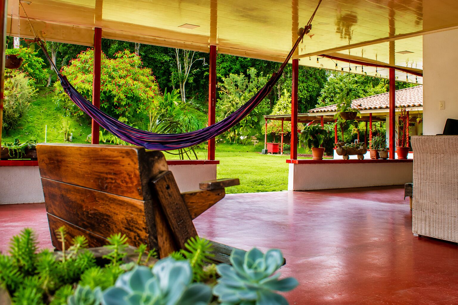 A hammock under a wooden structure with a view of a garden and a building with a tiled roof.