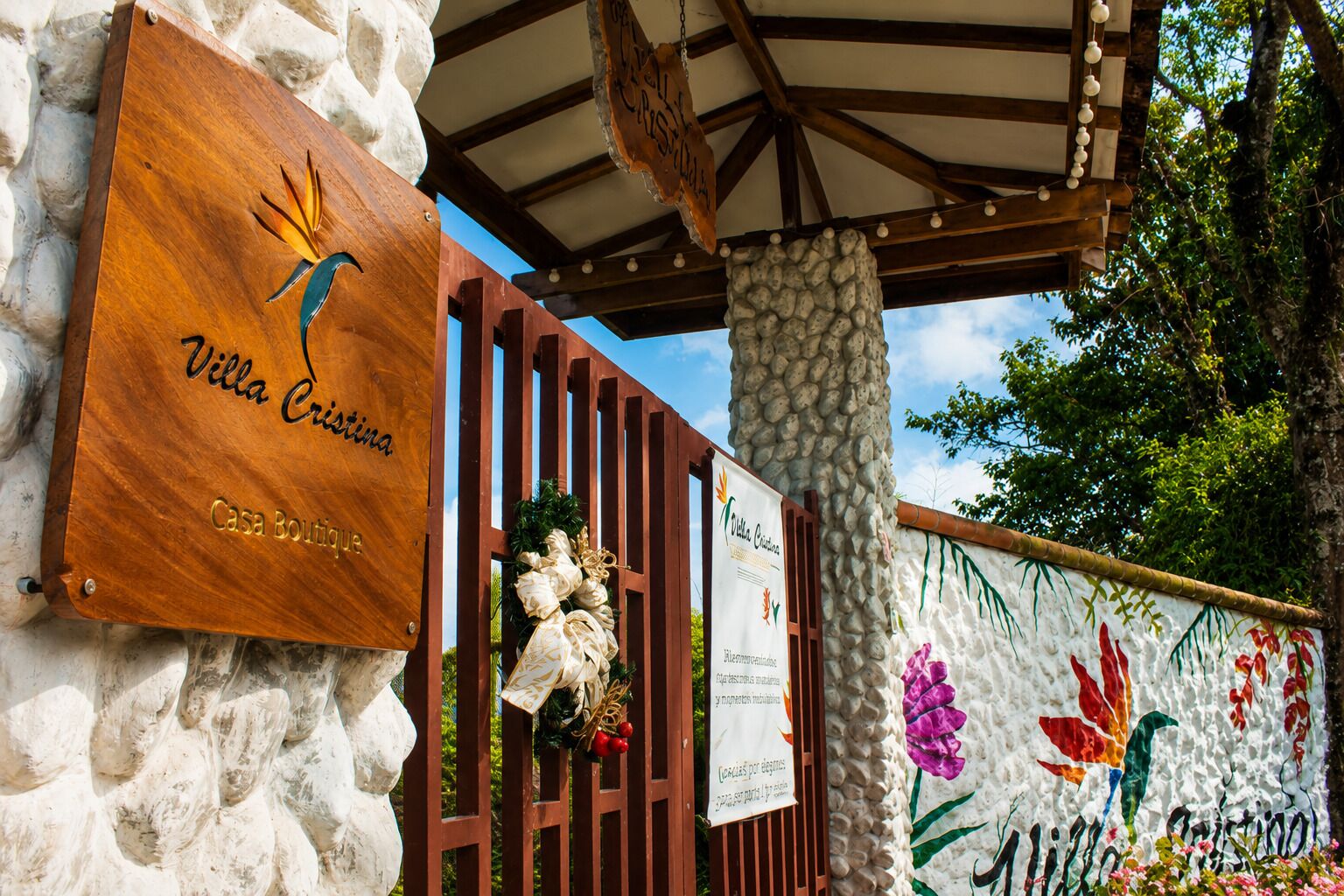 Un letrero de madera con 'Villa Cristina Casa Boutique' y un pilar de piedra con un muro decorativo con flores pintadas.