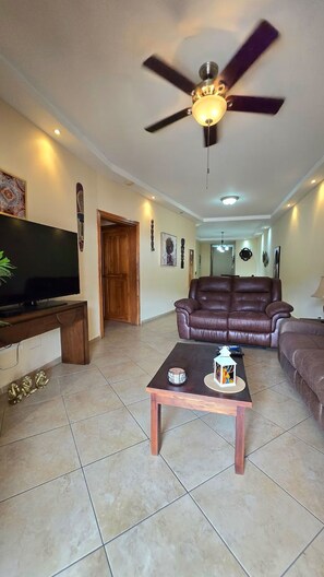A living room with a television, a ceiling fan, a leather sofa, and a coffee table. - Spacious Downtown Boquete 2BR/2BA Condo with Private Balcony (Bajo Boquete)