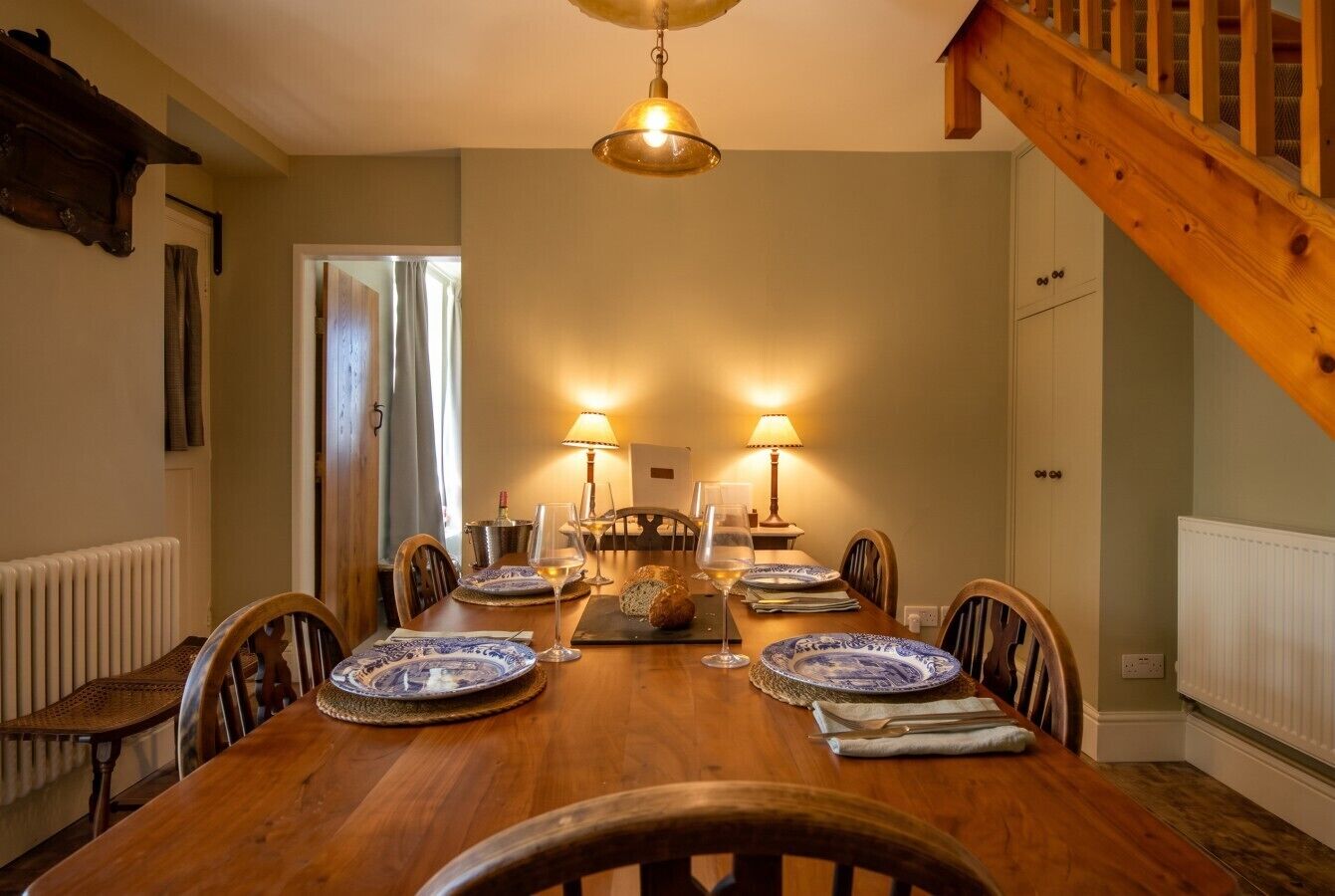 A dining room with a wooden table set for four, flanked by two lamps, a radiator, and a staircase.