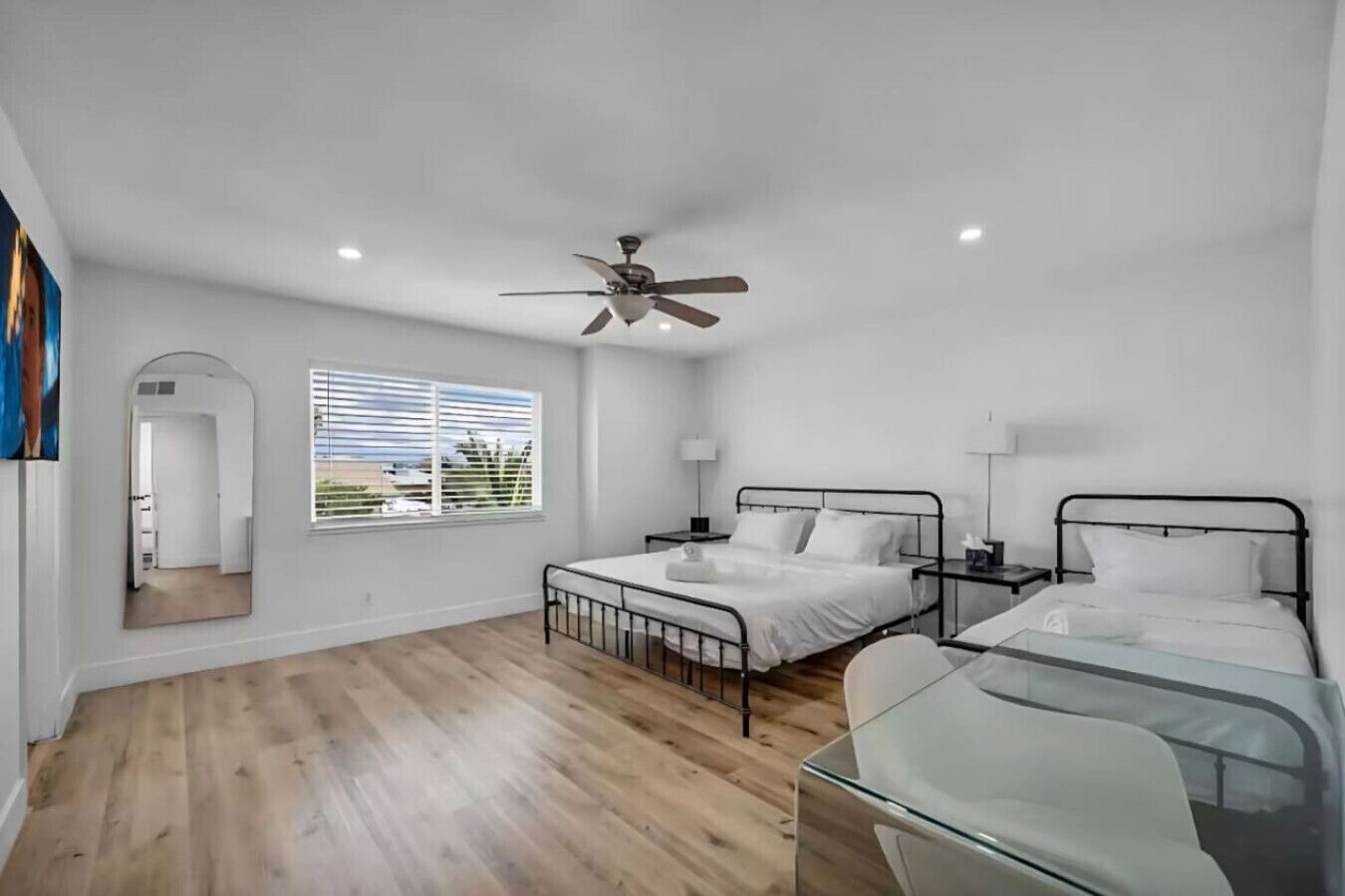 A hotel room with two beds, a ceiling fan, a window with blinds, a mirror, and a wooden floor.