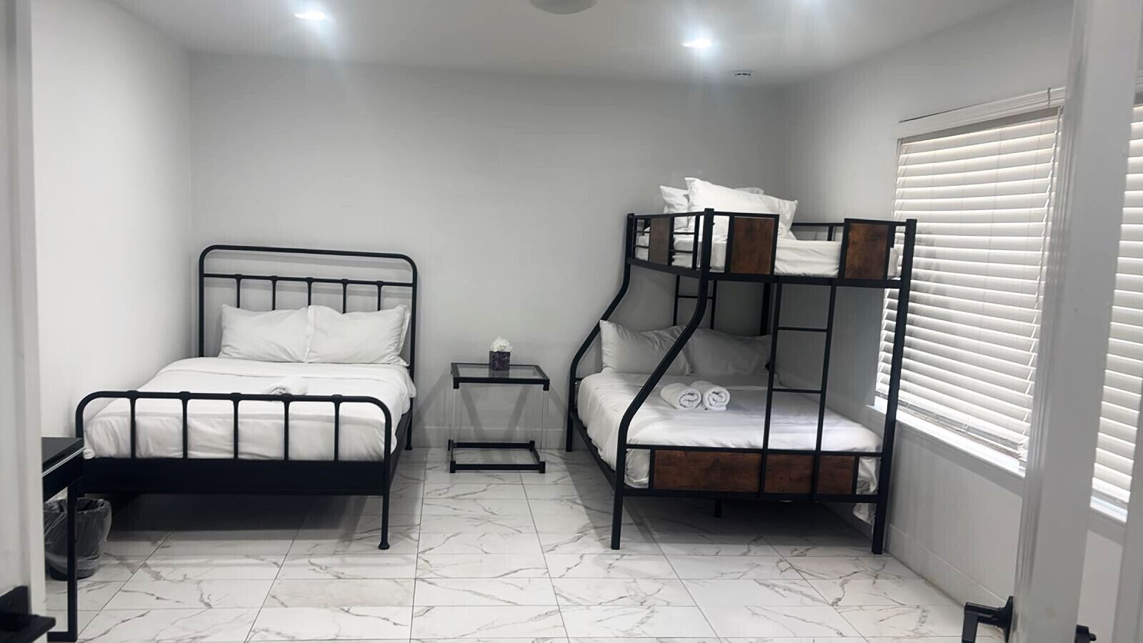 A room with two bunk beds, a small bedside table with a lamp, and a window with blinds.