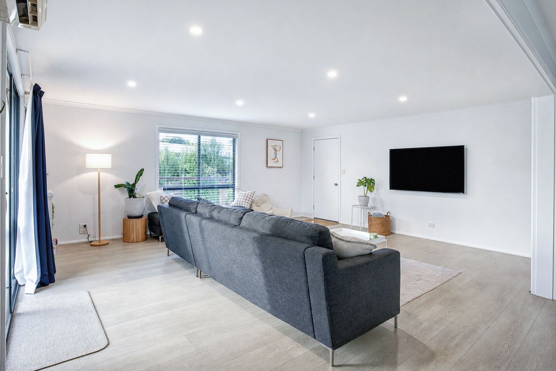 A modern living room with a grey sofa, a standing lamp, a flat-screen TV, and a large window with blinds.