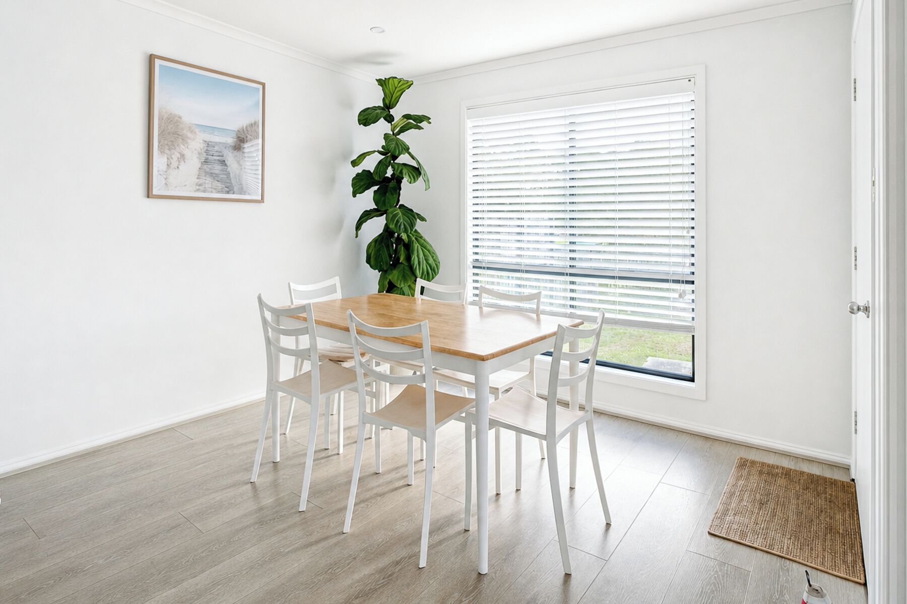 A dining area with a wooden table and white chairs, a large green plant, a window with blinds, and a framed picture on the wall.