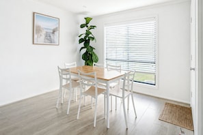 Ein Essbereich mit einem Holztisch und weißen Stühlen, einer großen Grünpflanze, einem Fenster mit Jalousien und einem gerahmten Bild an der Wand.