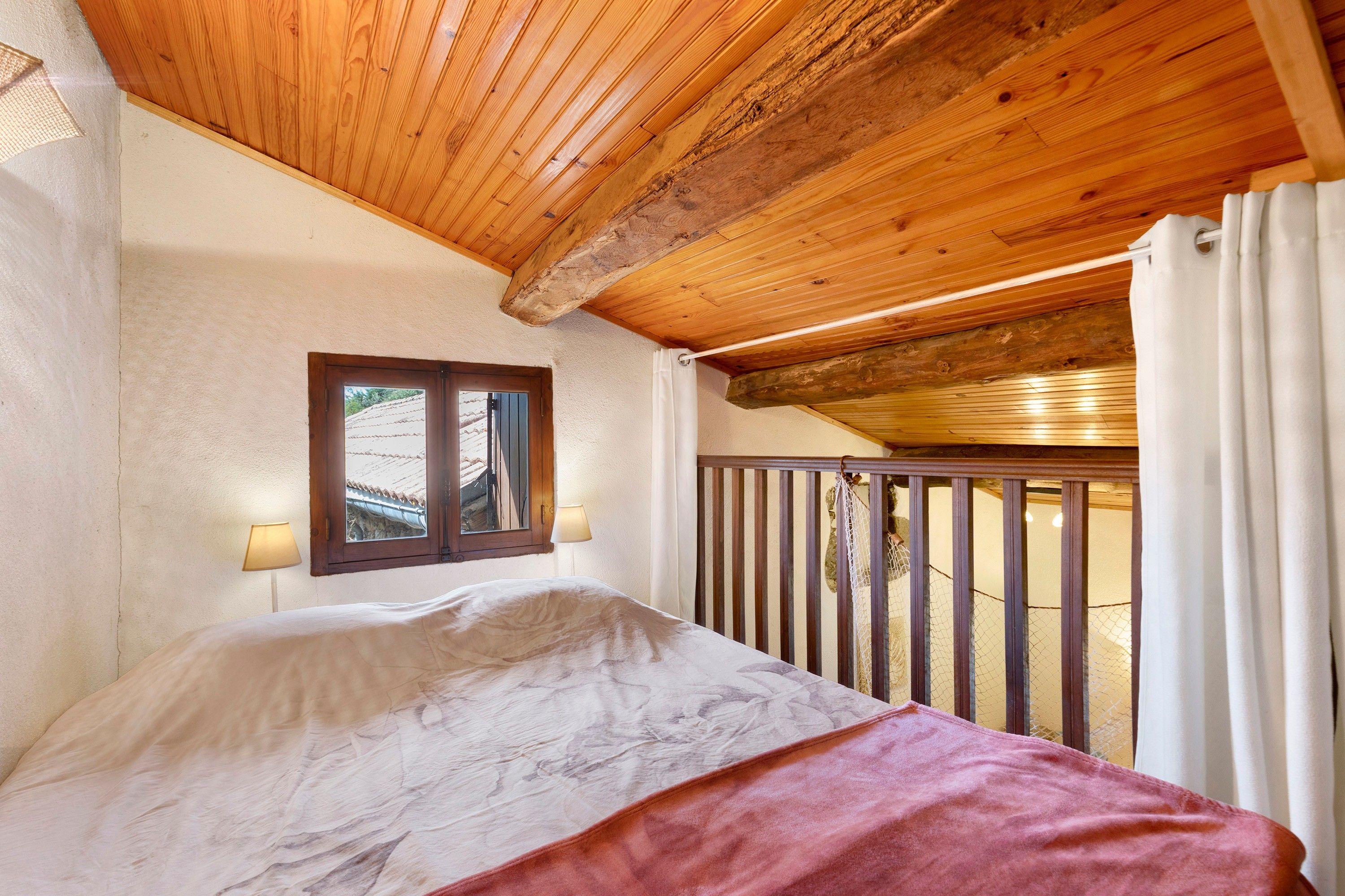 A bedroom with a wooden ceiling, a window with a view, a bed with a white and red blanket, and a balcony with a railing.