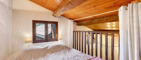 A bedroom with a wooden ceiling, a window with a view, a bed with a white and red blanket, and a balcony with a railing.