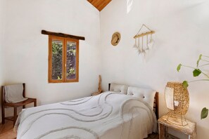 Un dormitorio con cama, ventana, una silla y una mesita de noche con lámpara.