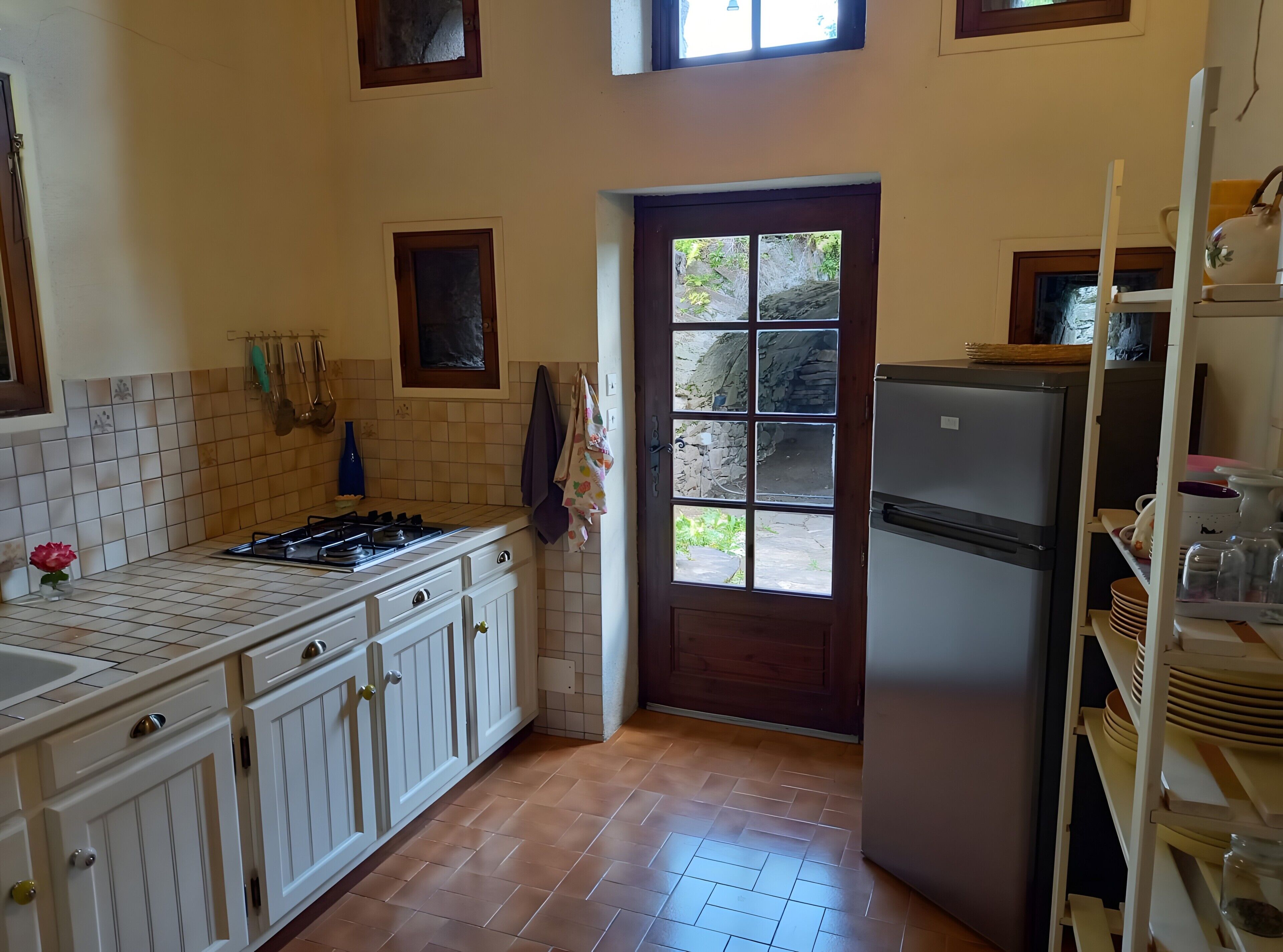Une cuisine avec des meubles blancs, une cuisinière à gaz, un réfrigérateur et une fenêtre donnant sur un espace extérieur rocheux.