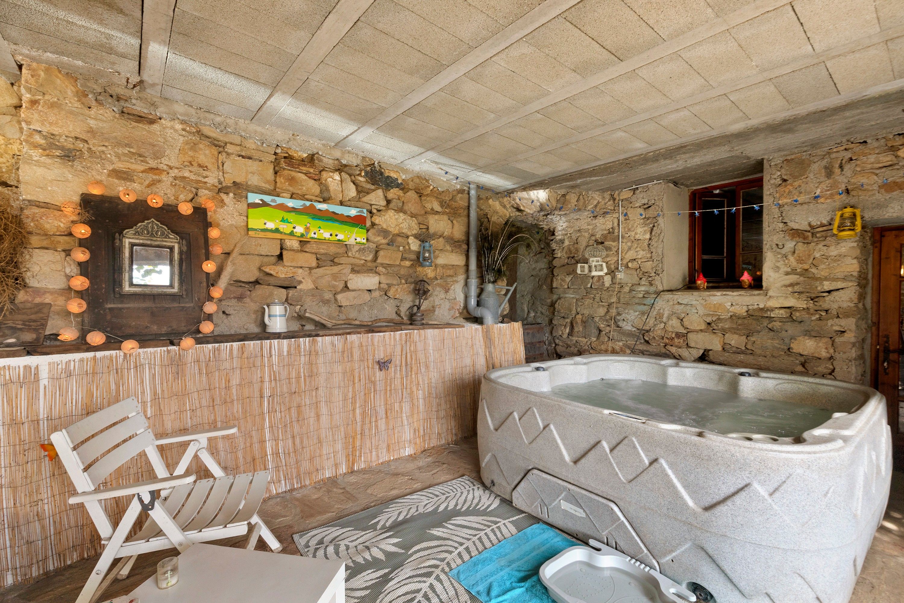 A stone-walled room with a wooden bench, a hot tub, and a rustic decor including a painting and lanterns.