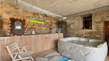 A stone-walled room with a wooden bench, a hot tub, and a rustic decor including a painting and lanterns.