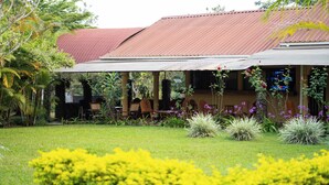 A building with a covered patio area, surrounded by lush greenery and flowering plants.