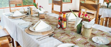 Una mesa de comedor dispuesta con platos blancos, cubiertos y servilletas, con un camino de mesa estampado y elementos decorativos como un jarrón con flores y botellas de condimentos.