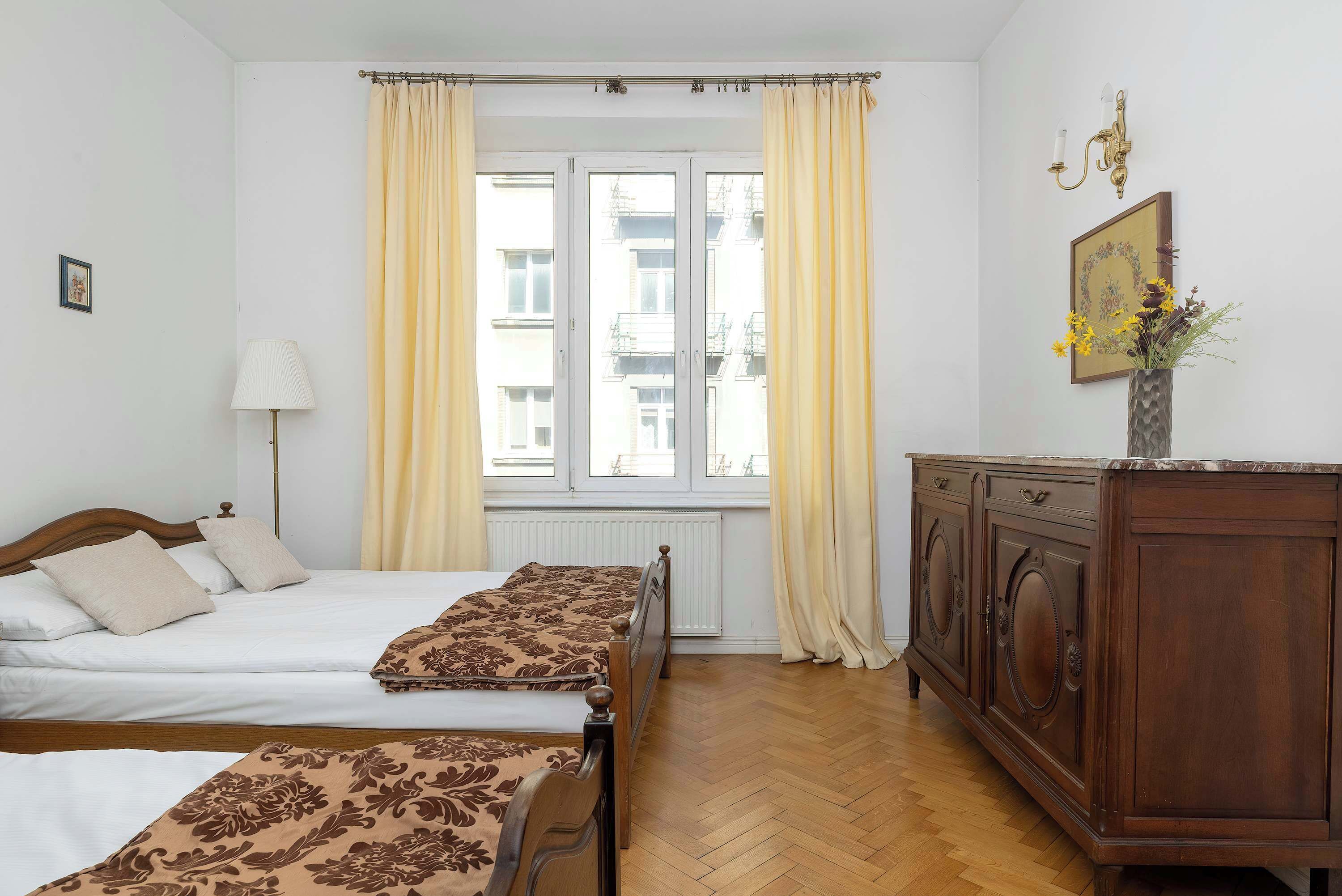 A hotel room with two beds, a wooden dresser, a radiator, and a window with curtains.
