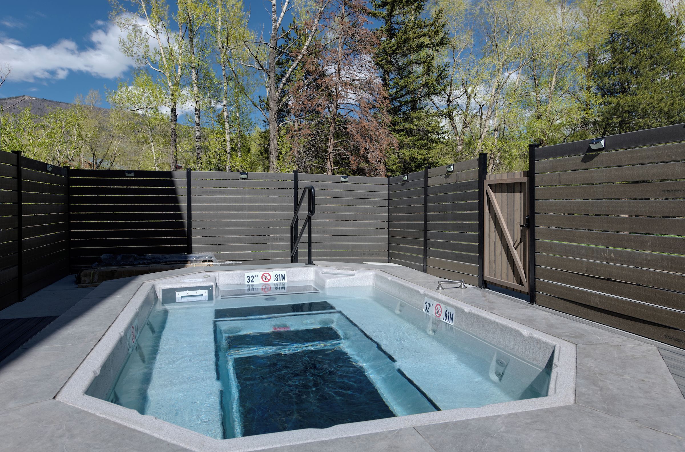 Un jacuzzi rectangulaire entouré d’une clôture en bois, avec en arrière-plan un paysage de forêts et de montagnes.