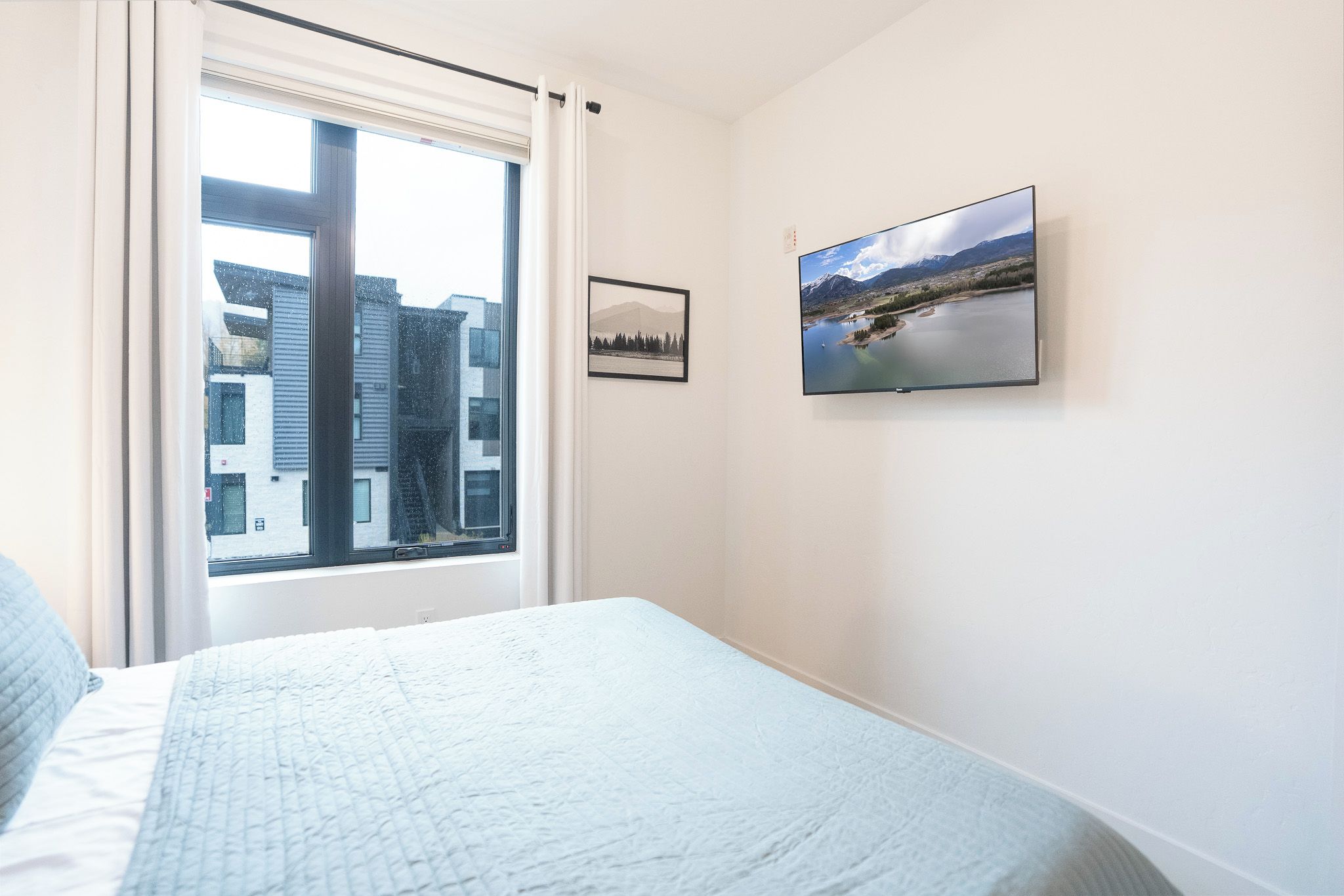 Une chambre à coucher avec un lit, une fenêtre donnant sur des immeubles et un téléviseur fixé au mur.