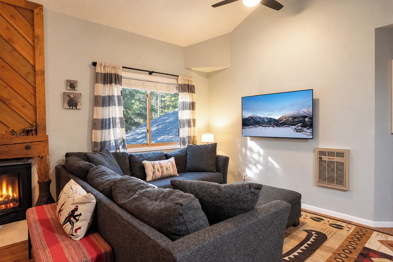 A cozy living room with a fireplace, a sectional sofa, a large window with curtains, a mounted TV, and a wall-mounted heater.