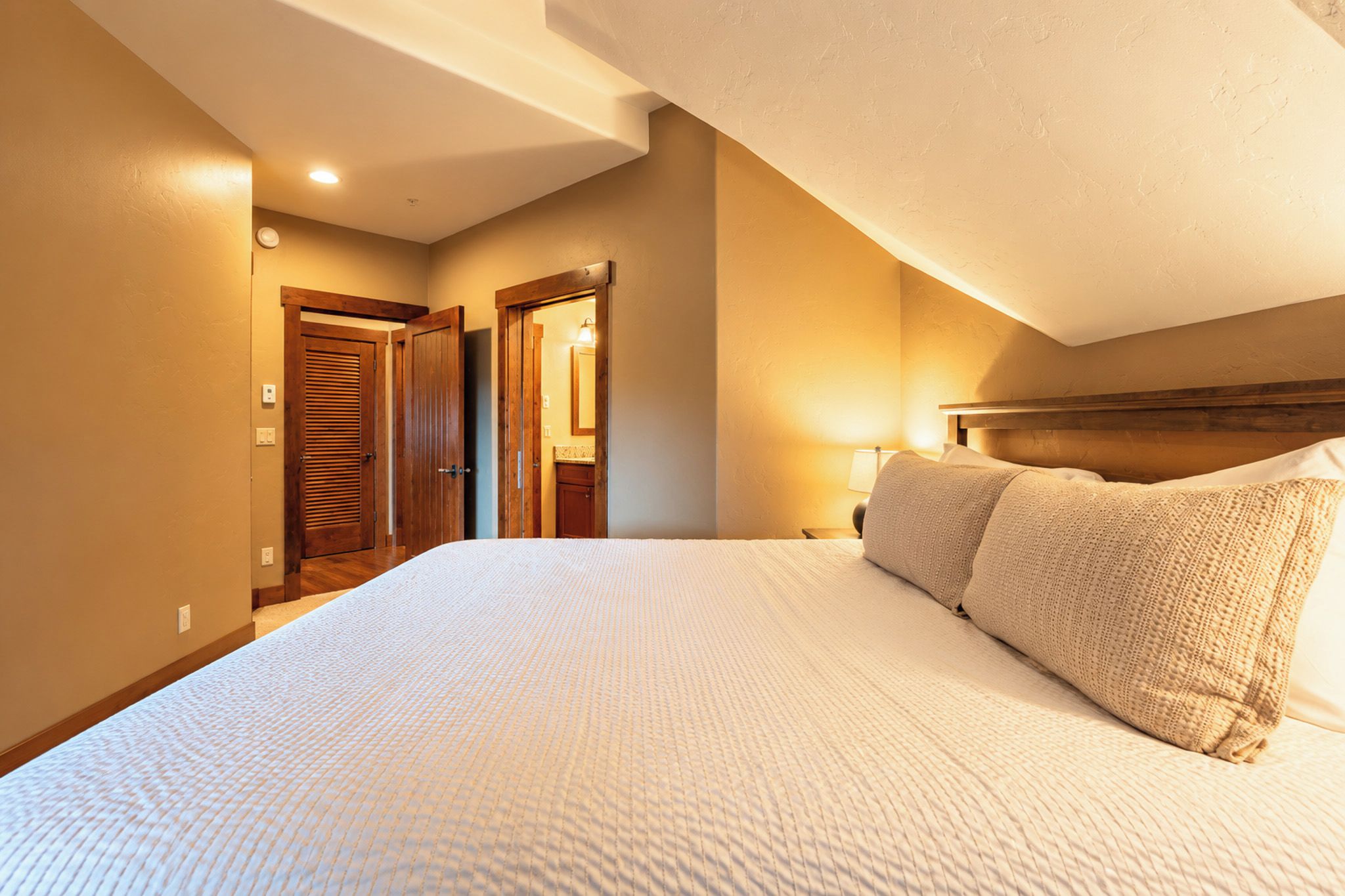A hotel room with a large bed, wooden doors, a nightstand, and a wall-mounted lamp.