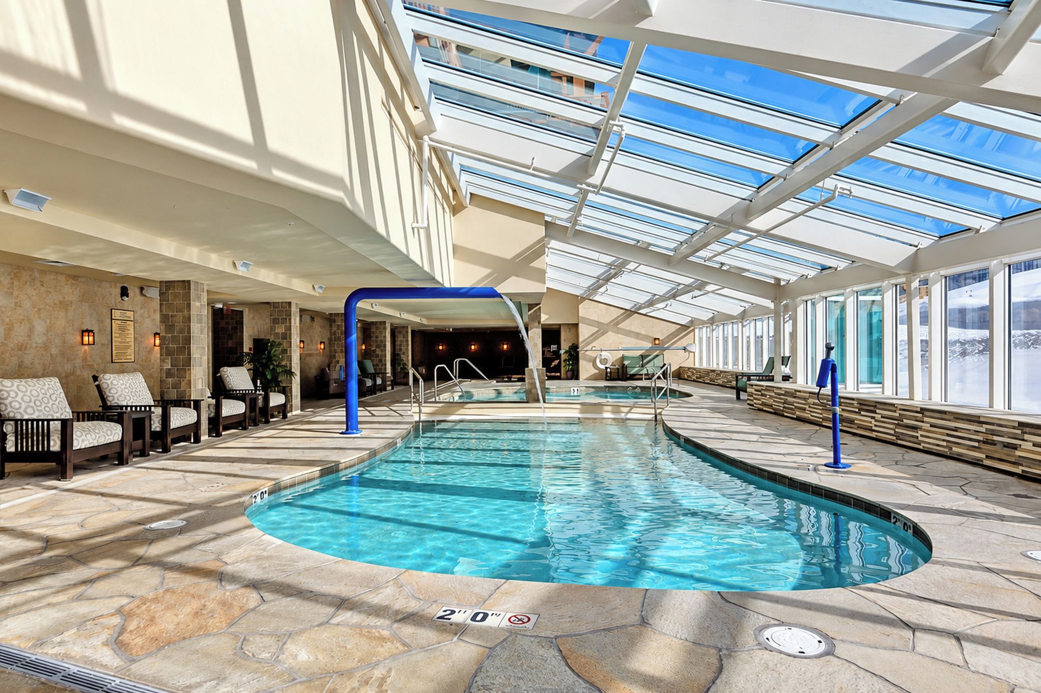 A large indoor swimming pool with a glass ceiling, stone flooring, and lounge chairs along the sides.