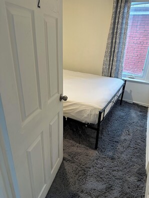 Une petite chambre à coucher avec un lit simple, une porte, une fenêtre avec des rideaux et un sol recouvert d’une moquette.