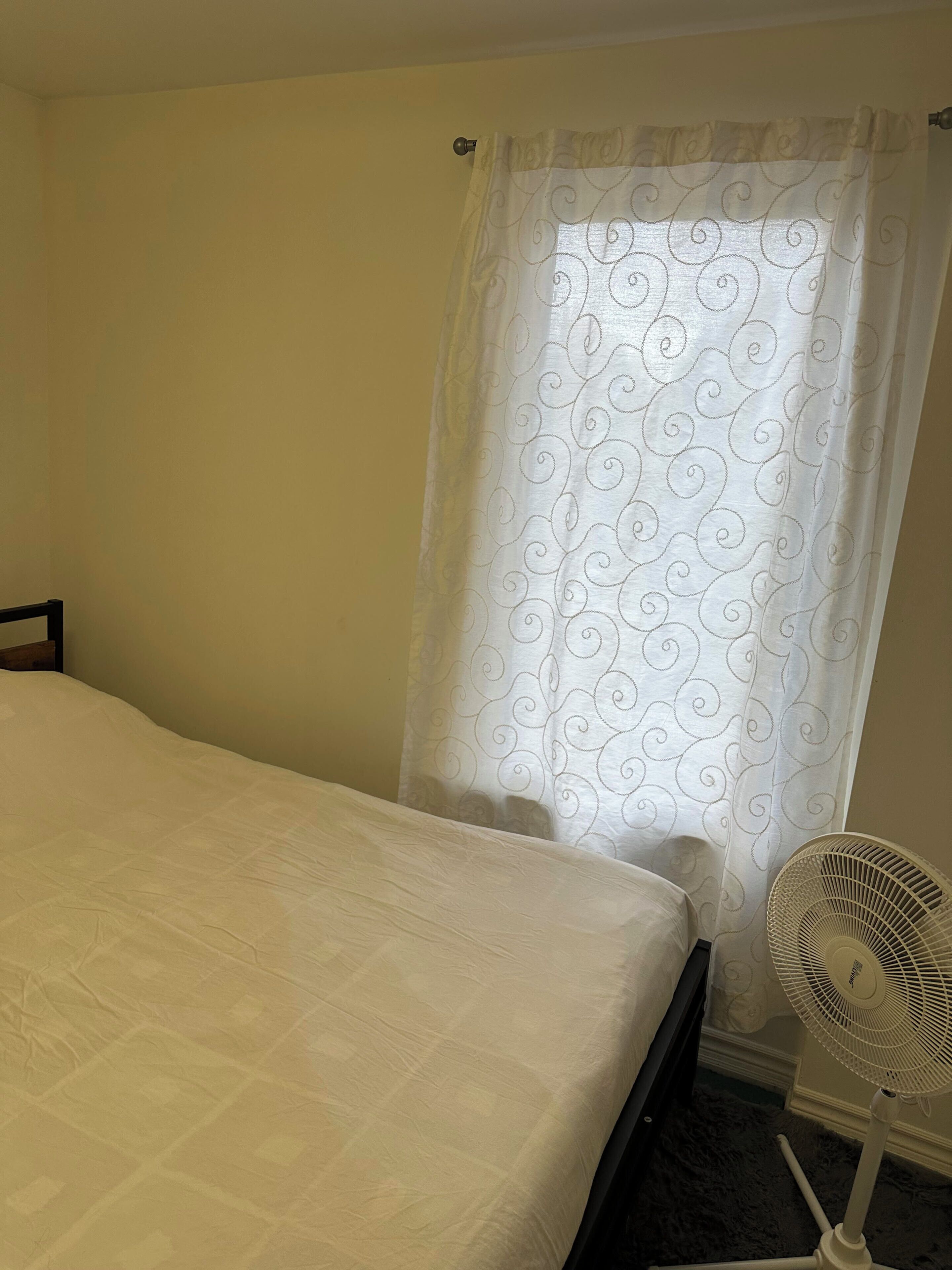 A bedroom with a bed, a white curtain with a swirl pattern, and a standing fan.