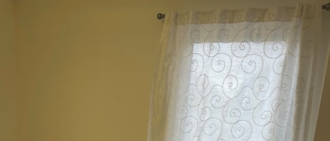 A bedroom with a bed, a white curtain with a swirl pattern, and a standing fan.