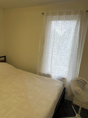 A bedroom with a bed, a white curtain with a swirl pattern, and a standing fan.
