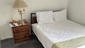 A bedroom with a bed, a nightstand with a lamp, a small wooden dresser, and a grey shaggy carpet. - Comfortable & Cozy Private Room in a Shared House (Room1) (Toronto)