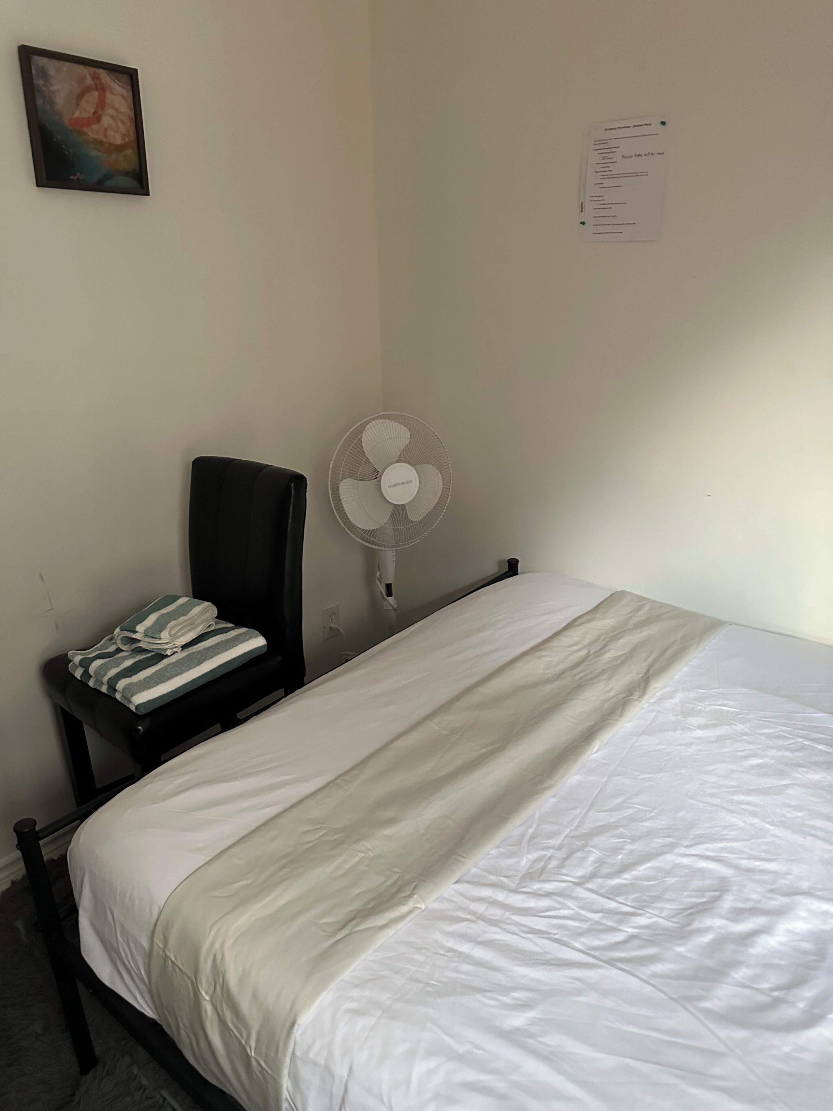 Ein schlichter Raum mit einem Bett, einem Stuhl, einem Standventilator und einem Gegenstand an der Wand.