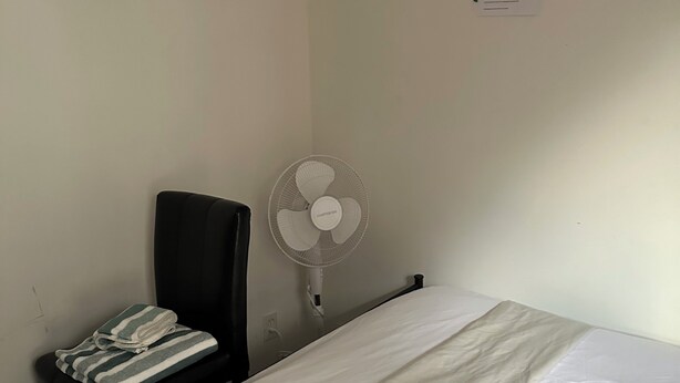 A simple room with a bed, a chair, a standing fan, and a wall-mounted item. - Comfortable & Cozy Private Room in a Shared House (Room1) (Toronto)