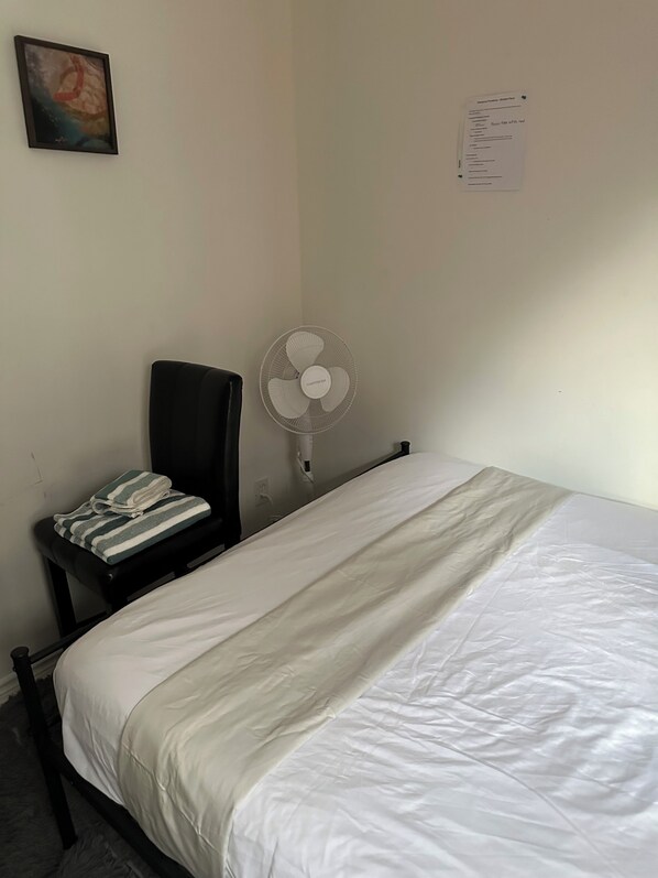 Une pièce simple, avec un lit, une chaise, un ventilateur sur pied et un objet fixé au mur.