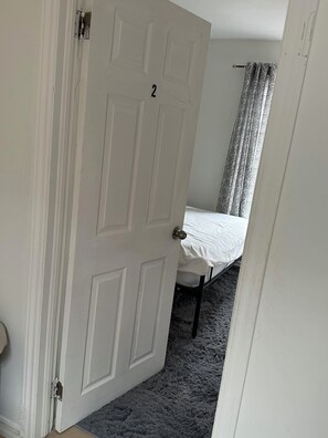 Une pièce avec une porte blanche portant le numéro 2, un lit avec un matelas blanc et un tapis gris.