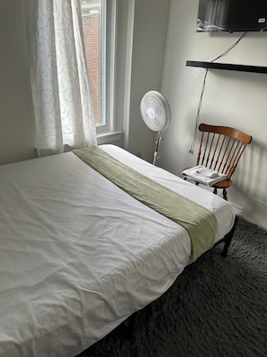 Un lit bien fait, avec un plaid vert, un ventilateur sur pied, une chaise en bois et un téléviseur fixé au mur.