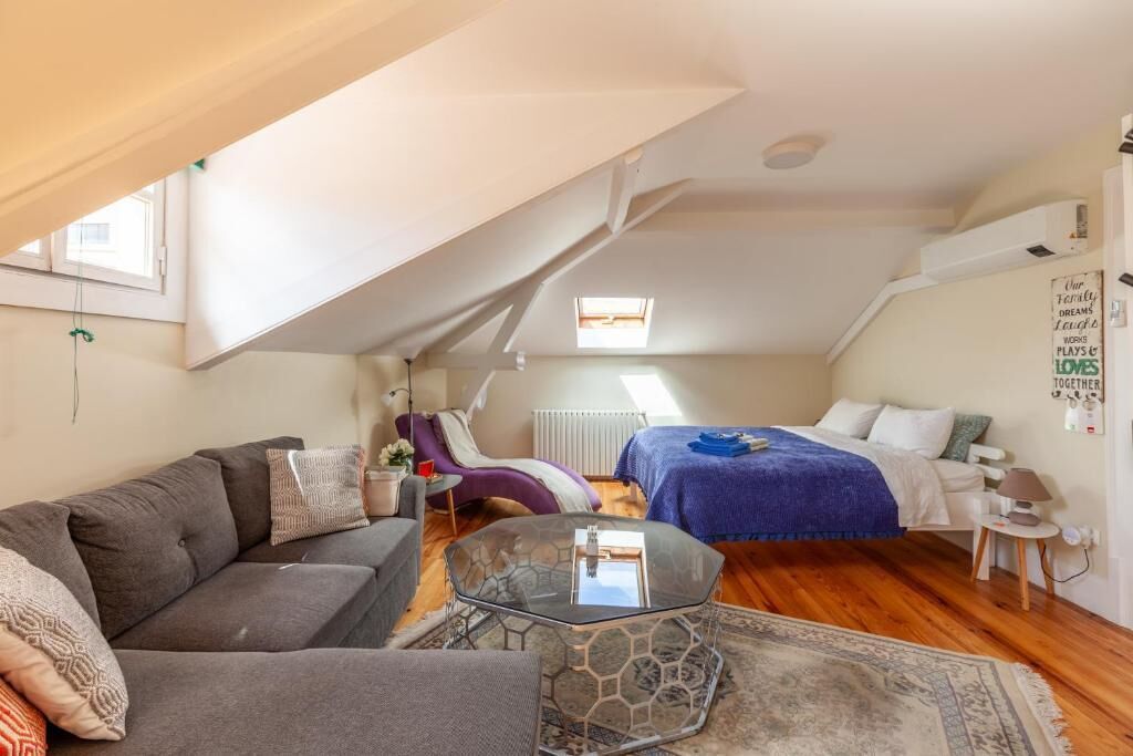 Een kleine kamer met een bank, een glazen bijzettafeltje, een bed met een blauwe deken, en een klein nachtkastje.