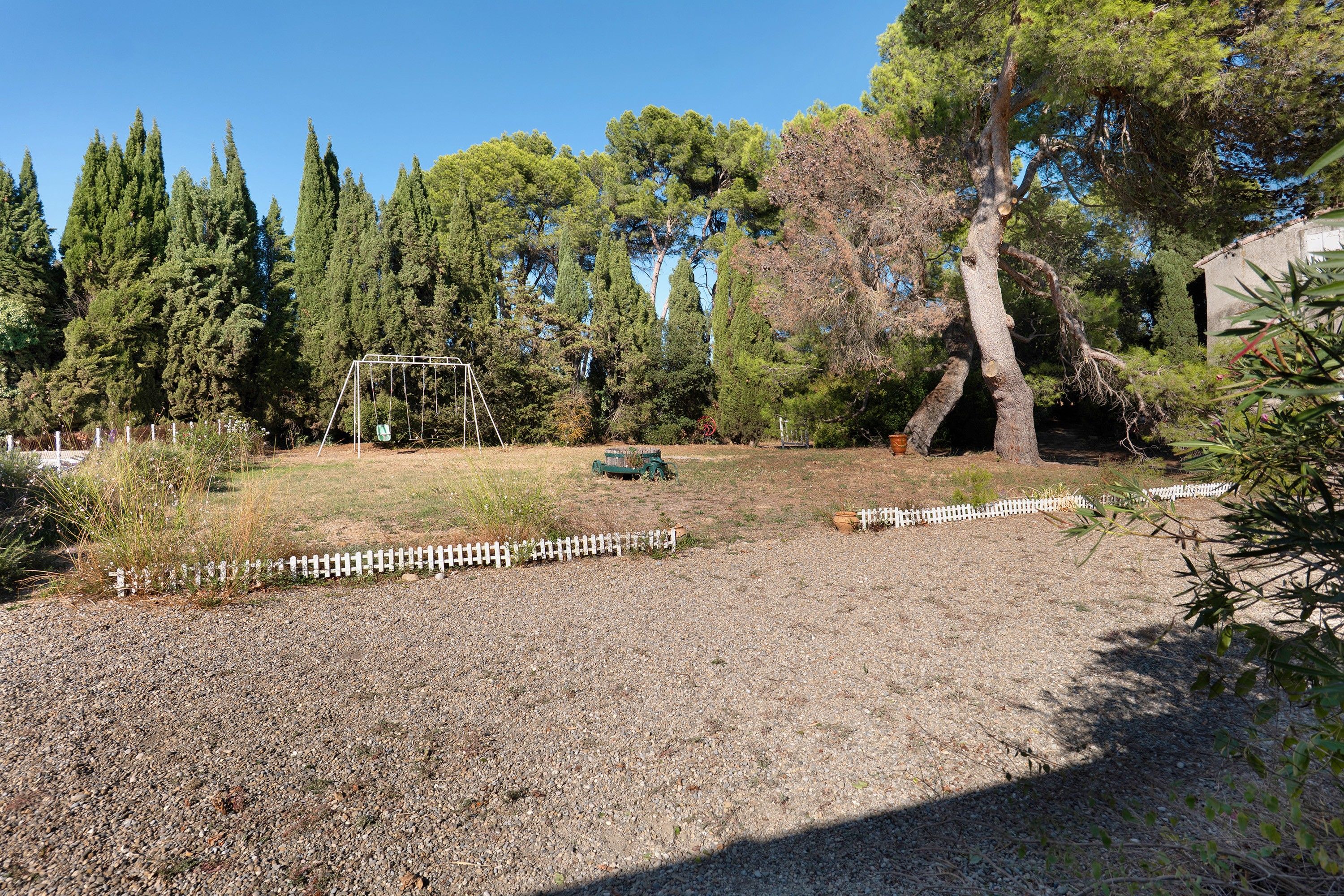 A gravel area with a white picket fence, a swing set, and a large tree.
