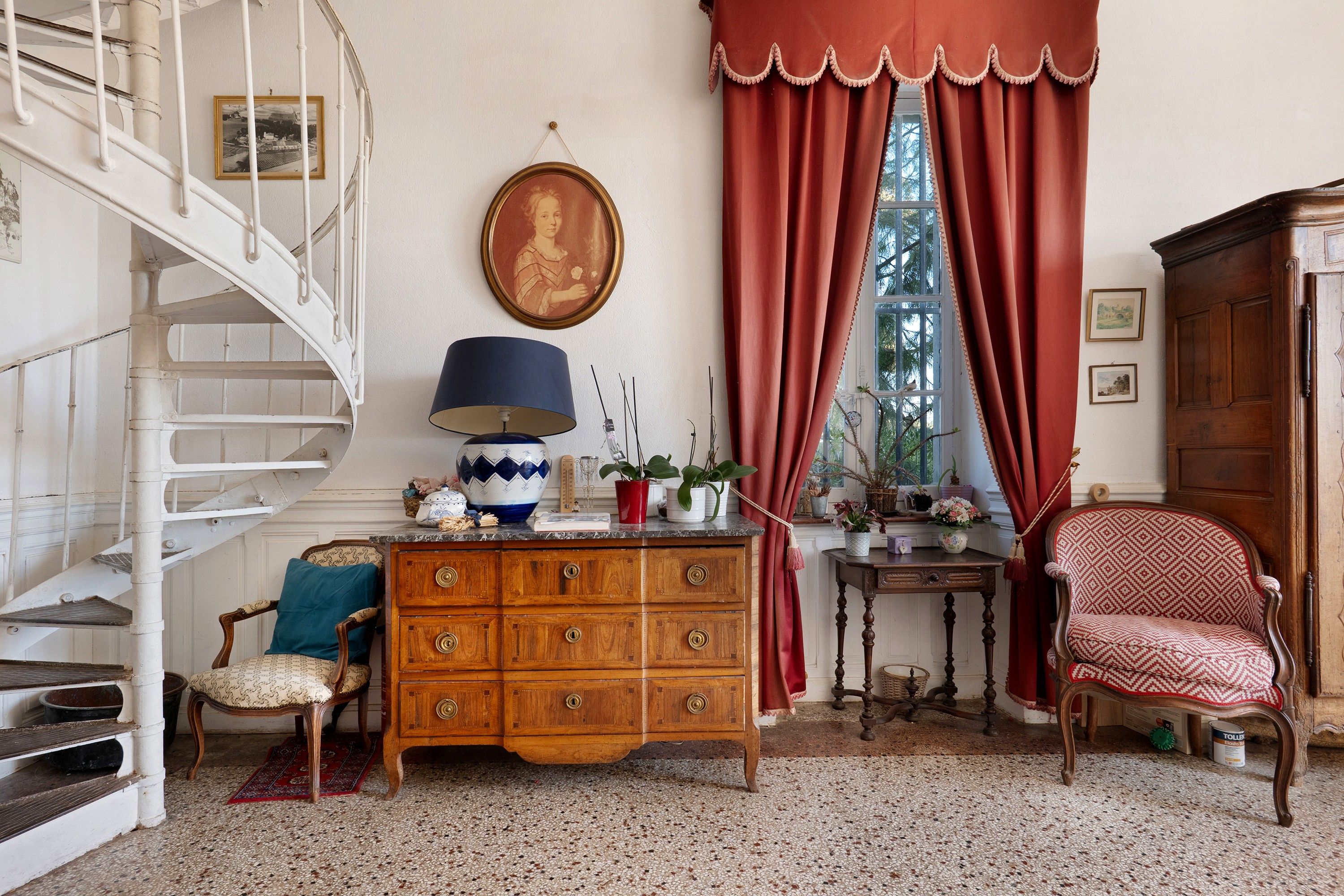 Une pièce avec une commode en bois, une chaise, une table avec une lampe, un rideau rouge et un escalier.