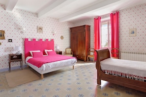 A bedroom with a large bed, a smaller bed, a wooden wardrobe, a chair, and a window with curtains.