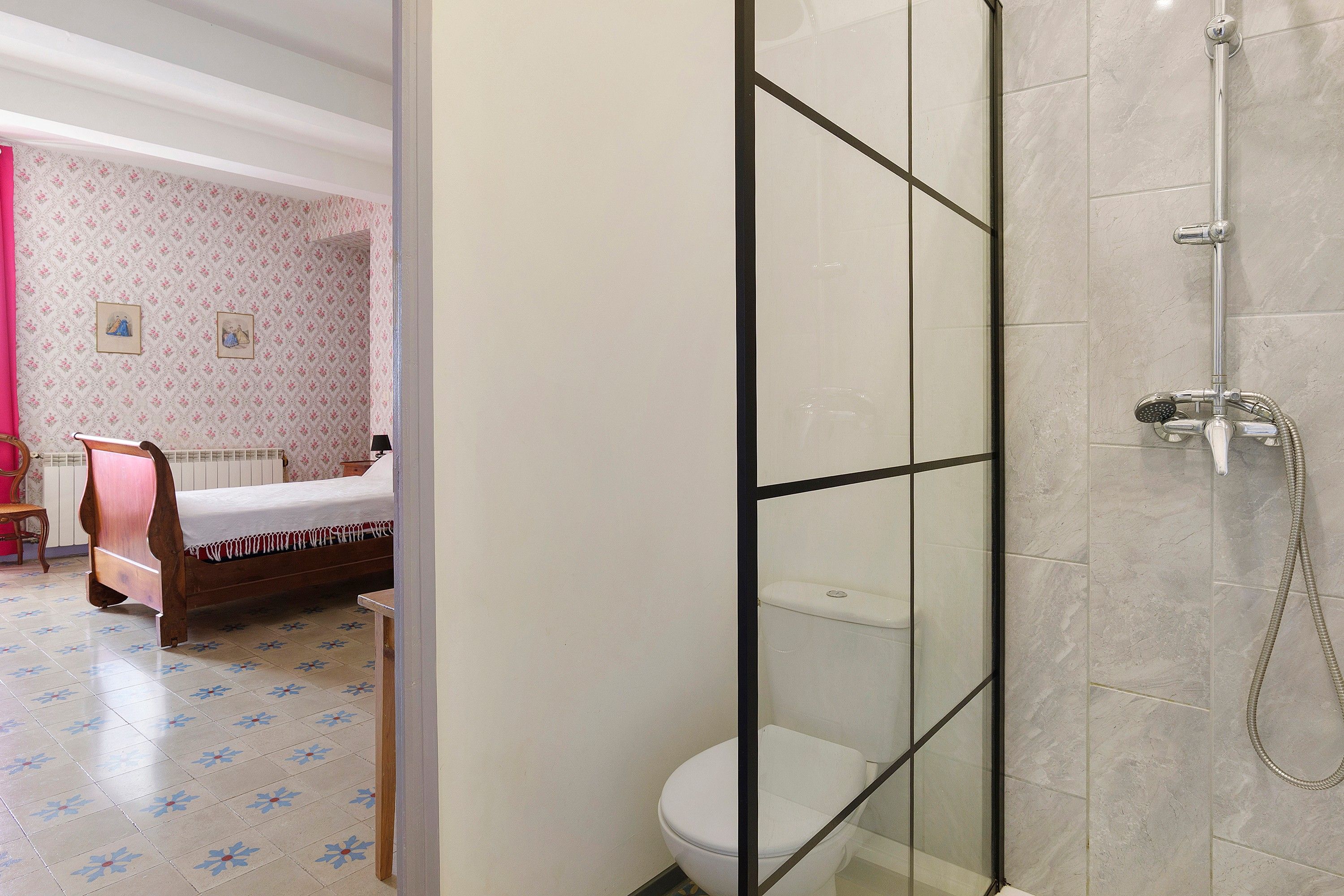 A bathroom with a glass shower enclosure, a toilet, and a bed with floral-patterned wallpaper in the adjacent room.
