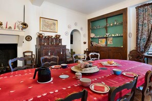 Une salle à manger avec une nappe rouge, des chaises en bois, une cheminée et un mur vert agrémenté d’étagères.