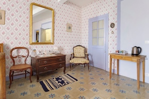 A room with a wooden dresser, a chair, a table with a kettle, a door, and a mirror on the wall.