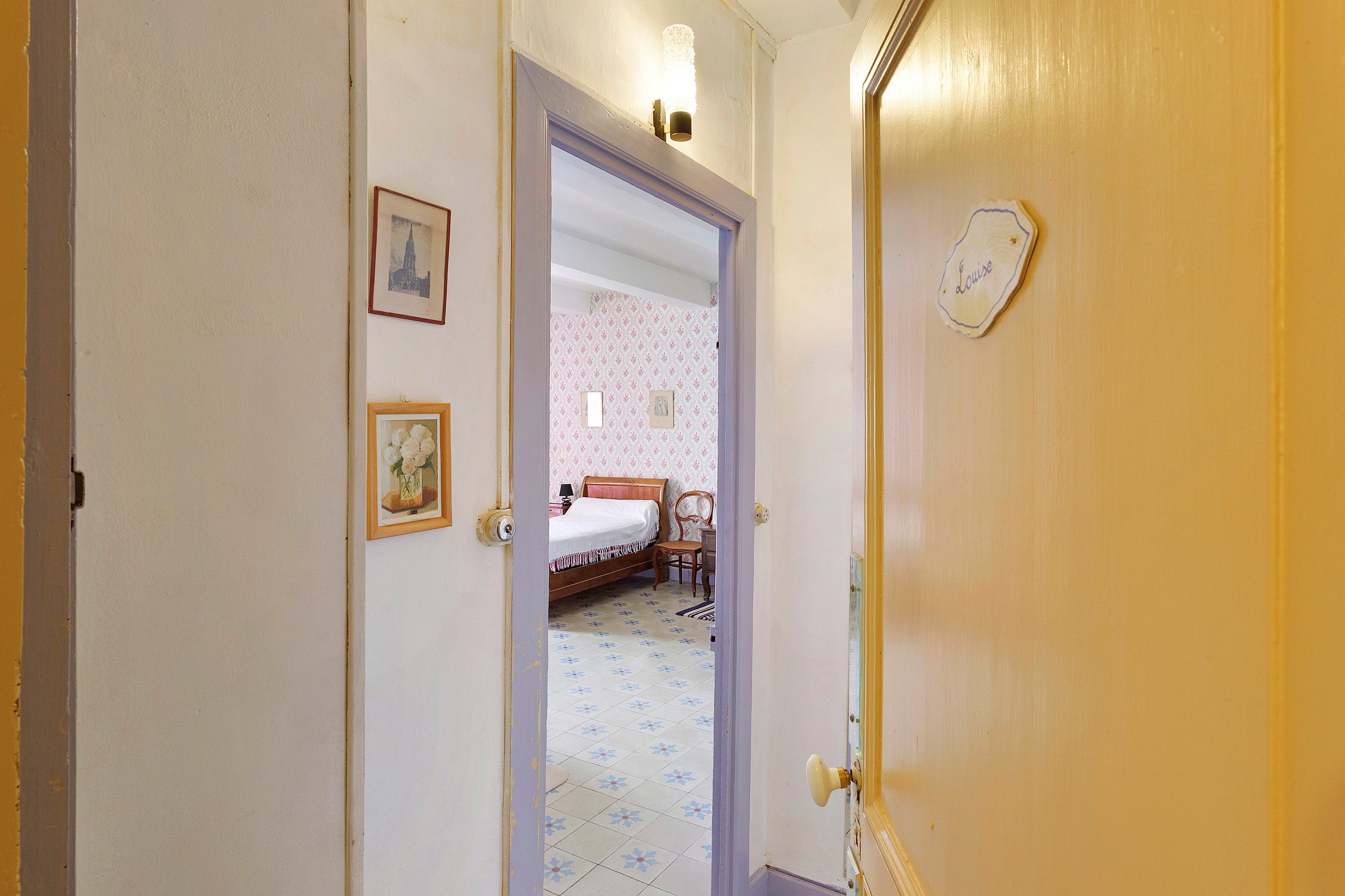 Een gang die uitkomt in een slaapkamer met een bed, een stoel en een klein tafeltje.