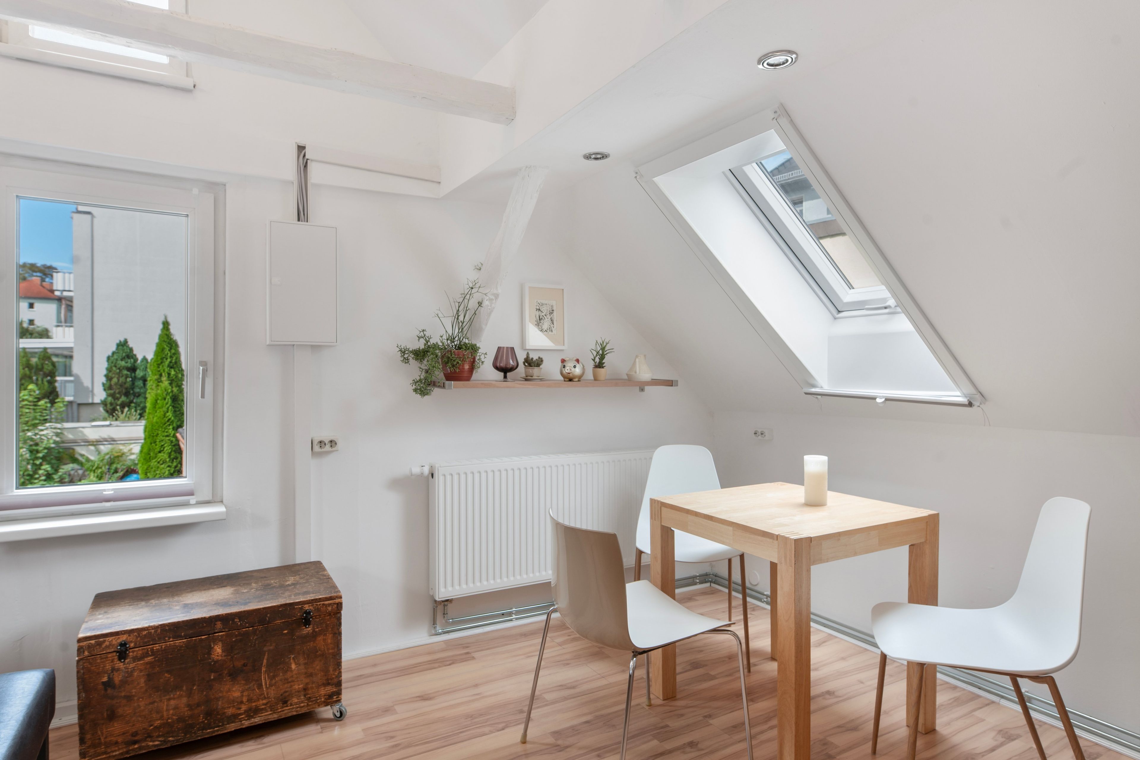 Una habitación moderna y bien iluminada, con techo inclinado, tragaluz, radiador blanco, mesa de madera con sillas y un aparador rústico.