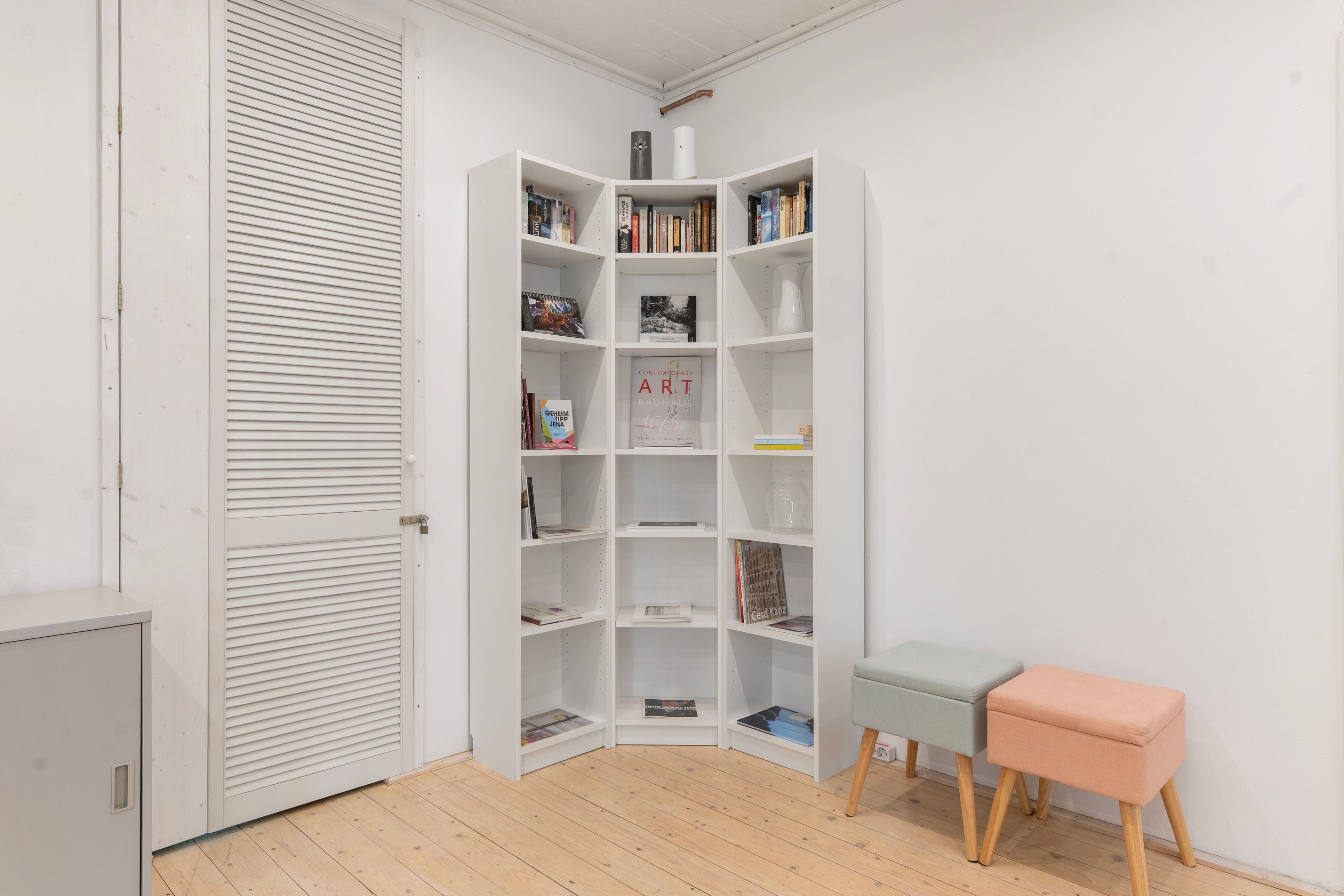 Una habitación de esquina con una estantería blanca, un banco gris y una puerta cerrada con persianas horizontales.