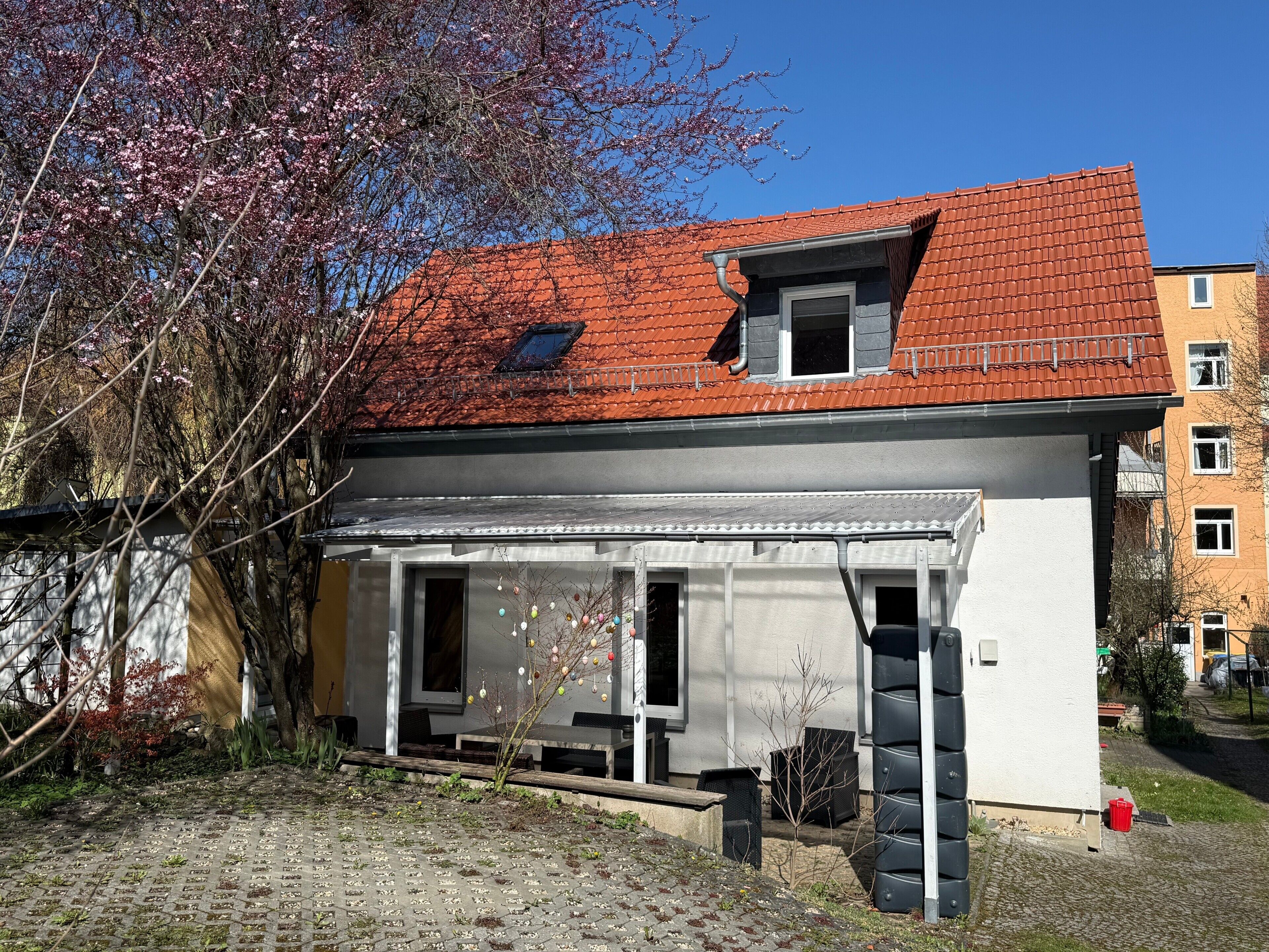 Ein zweistöckiges Haus mit rotem Ziegeldach, einem kleinen Balkon und einem Garten mit Baum und Bank.